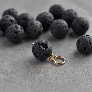 May include: Close-up of black lava rock beads with a gold-coloured charm loop. The porous, round beads are scattered on a grey surface. The image shows the detail of the lava rock texture. The text at the bottom says "(For 1 finished charm. Stone randomly selected. Enlarged to show detail)".