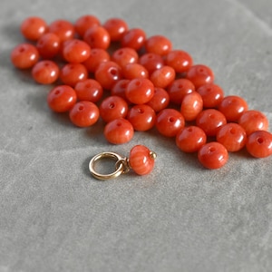 May include: A collection of small, round, vibrant orange beads scattered on a gray surface. A single charm with a gold-colored ring and an orange bead is also present. The beads appear to be made of coral.