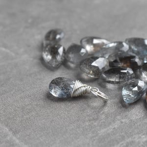 May include: A collection of faceted, teardrop-shaped gemstones in shades of gray and blue. One stone is wire-wrapped with a silver-colored metal loop. The text at the bottom reads: "(For 1 finished charm. Stone randomly selected. Enlarged to show detail)"