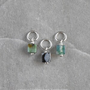 May include: Three gemstone charms with silver hoops. Each charm has a small, square-cut stone in shades of green and black. The charms are displayed on a grey surface.