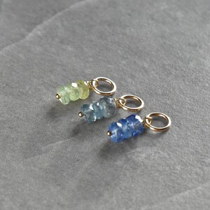 May include: Three gold charms with different colored gemstone beads. The charms are in a row from left to right: light green, light blue, and dark blue.