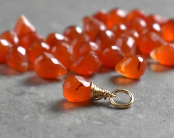 May include: A close-up of a carnelian gemstone charm, wrapped in gold wire and attached to a gold jump ring. The charm is orange and has a faceted shape. There are many other carnelian gemstones in the background.