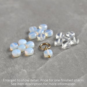 May include: A close-up of a charm made with a white opal gemstone and a gold jump ring. The charm is surrounded by loose white opal and clear glass beads.