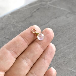 Puede incluir: Un peque&ntilde;o amuleto de piedra lunar iridiscente con un envoltorio de alambre dorado y un anillo de salto dorado.