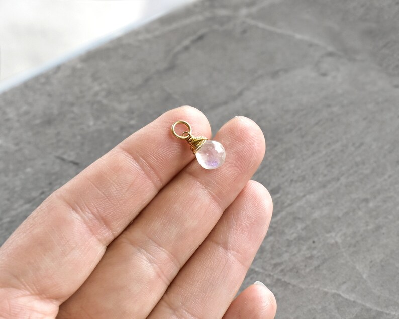Puede incluir: Un peque&ntilde;o amuleto de piedra lunar iridiscente con un envoltorio de alambre dorado y un anillo de salto dorado.