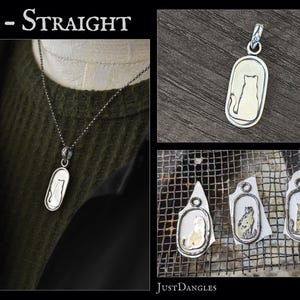 May include: A silver pendant necklace featuring a cat silhouette within an oval frame. The pendant hangs from a delicate chain. The image also shows the pendant on a dark green sweater and some work in progress.