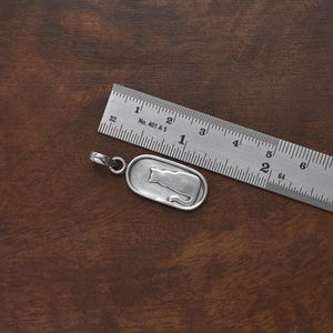 May include: A silver pendant with a cat silhouette design, next to a metal ruler. The pendant is approximately 1 inch long. The ruler shows measurements in inches. The background is a dark brown wooden surface.
