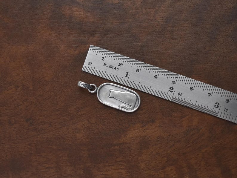 May include: A silver pendant with a cat silhouette design, next to a metal ruler. The pendant is approximately 1 inch long. The ruler shows measurements in inches. The background is a dark brown wooden surface.