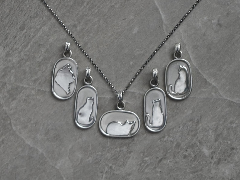May include: A silver necklace featuring a chain and six pendants. The pendants are rectangular and oval-shaped, each with a cat silhouette design. The cat pendants are in various poses, including sitting and lying down. The necklace is displayed on a gray surface.