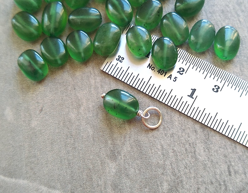 May include: A close-up of a green gemstone pendant with a silver bail. The pendant is about 1 inch long and is sitting on a ruler. The ruler is marked with inches and centimeters. The pendant is a popular choice for jewelry making.