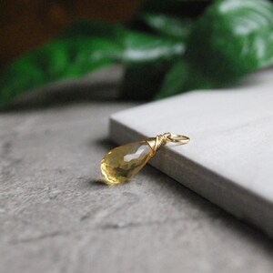 Puede incluir: Un colgante de piedra preciosa amarilla en forma de l&aacute;grima con un envoltorio de alambre dorado. El colgante est&aacute; sobre una superficie blanca.