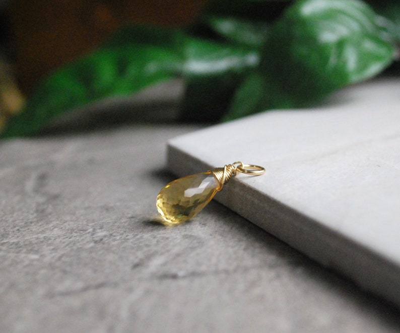 Puede incluir: Un colgante de piedra preciosa amarilla en forma de l&aacute;grima con un envoltorio de alambre dorado. El colgante est&aacute; sobre una superficie blanca.
