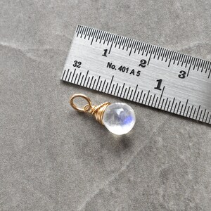 Puede incluir: Un peque&ntilde;o colgante de piedra lunar transparente e iridiscente envuelto en alambre dorado. El colgante tiene aproximadamente 1,25 cm de di&aacute;metro y se muestra junto a una regla para referencia de tama&ntilde;o.