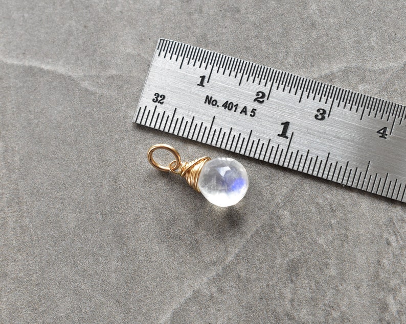 Puede incluir: Un peque&ntilde;o colgante de piedra lunar transparente e iridiscente envuelto en alambre dorado. El colgante tiene aproximadamente 1,25 cm de di&aacute;metro y se muestra junto a una regla para referencia de tama&ntilde;o.