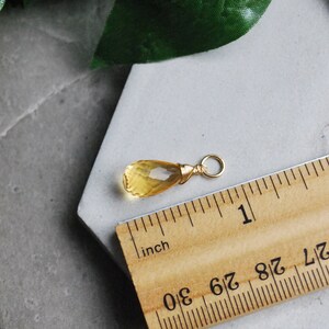 Puede incluir: Un colgante de citrino amarillo en forma de l&aacute;grima con un envoltorio de alambre dorado y un anillo de salto dorado. El colgante mide aproximadamente 2,5 cm de largo y se muestra junto a una regla.