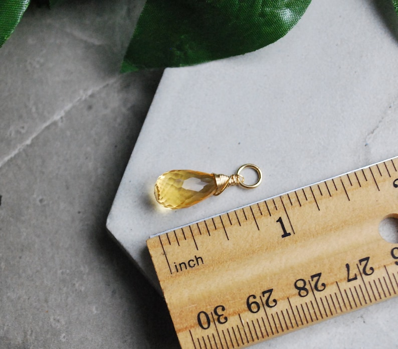 Puede incluir: Un colgante de citrino amarillo en forma de l&aacute;grima con un envoltorio de alambre dorado y un anillo de salto dorado. El colgante mide aproximadamente 2,5 cm de largo y se muestra junto a una regla.