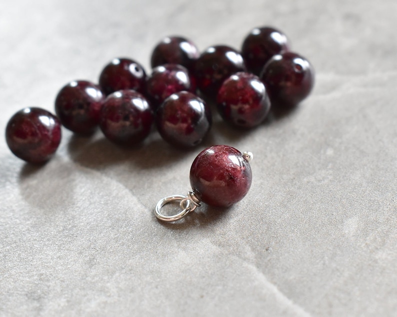 May include: A group of deep red gemstone beads, some of which are strung on a silver chain. The beads are round and smooth, and they have a slightly iridescent sheen.