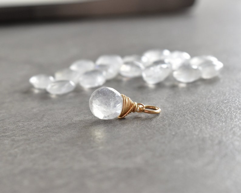 May include: Close-up of a gold wire-wrapped pendant featuring a cloudy, translucent white gemstone. The pendant's simple design highlights the natural beauty of the stone.  A scattering of similar, smaller, tumbled white stones is visible in the background, suggesting a collection or related product line. The overall aesthetic is minimalist and elegant, suitable for jewelry lovers seeking delicate, natural pieces.