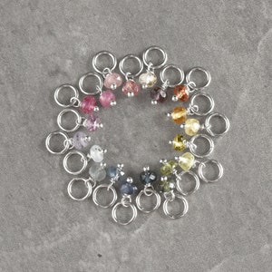 Op de afbeelding: Een cirkel van 18 zilveren springringen met gekleurde edelsteen kralen eraan bevestigd. De kralen zijn gerangschikt in een regenboogpatroon.