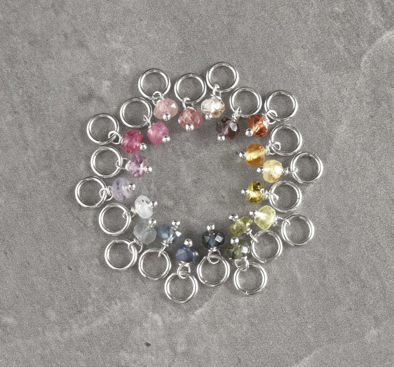 Op de afbeelding: Een cirkel van 18 zilveren springringen met gekleurde edelsteen kralen eraan bevestigd. De kralen zijn gerangschikt in een regenboogpatroon.
