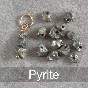 K&ouml;nnte beinhalten: Eine Sammlung roher Pyrit-Steine, von denen einige auf einer goldenen Kette mit einer kleinen Schlaufe oben aufgef&auml;delt sind. Die Steine sind in verschiedenen Graut&ouml;nen gehalten und haben eine raue, nat&uuml;rliche Textur. Das Wort "Pyrite" ist in wei&szlig;er Schrift unter den Steinen geschrieben.