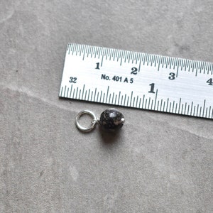 May include: A black and white speckled bead with a silver jump ring, measured against a ruler. The bead is approximately 1.2 cm in diameter.