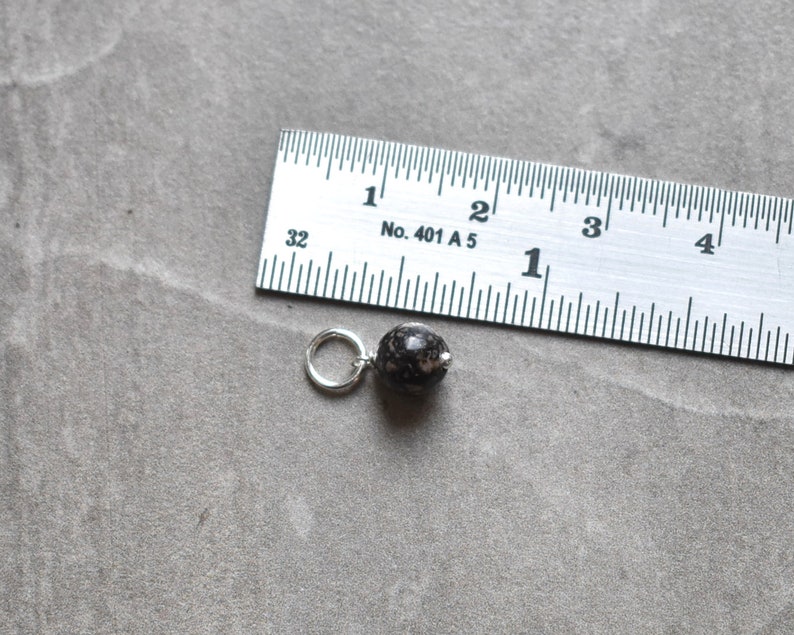May include: A black and white speckled bead with a silver jump ring, measured against a ruler. The bead is approximately 1.2 cm in diameter.