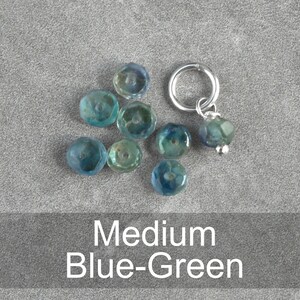 Op de afbeelding: Een set blauwe-groene glazen kralen met een zilveren kraal en ringbedel. De kralen zijn gefacetteerd en hebben een licht iriserende afwerking.