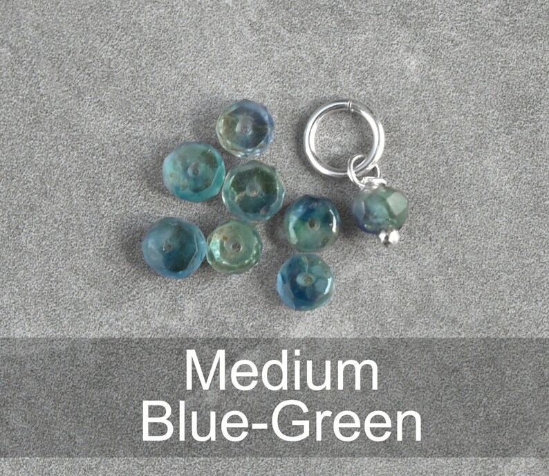 Op de afbeelding: Een set blauwe-groene glazen kralen met een zilveren kraal en ringbedel. De kralen zijn gefacetteerd en hebben een licht iriserende afwerking.