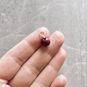 May include: A small, round, dark red gemstone bead with a silver loop attached. The bead is being held by a hand.