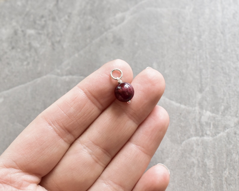 May include: A small, round, dark red gemstone bead with a silver loop attached. The bead is being held by a hand.