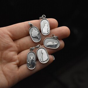 May include: A collection of silver cat pendant necklaces in various poses. Each pendant features a cat silhouette within an oval or rectangular frame. The pendants are held in a hand against a dark background.