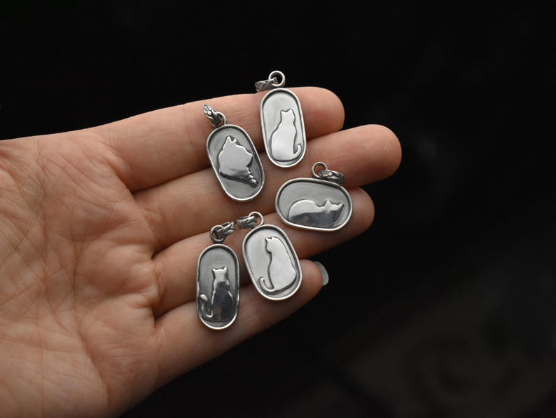 May include: A collection of silver cat pendant necklaces in various poses. Each pendant features a cat silhouette within an oval or rectangular frame. The pendants are held in a hand against a dark background.