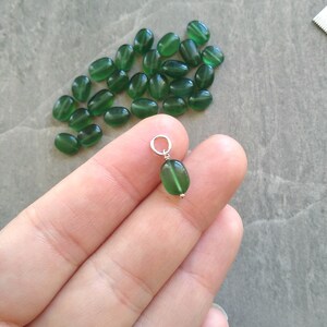 May include: A close-up of a green oval-shaped pendant with a silver bail. The pendant is held in a hand, and there are many other green oval-shaped beads in the background.