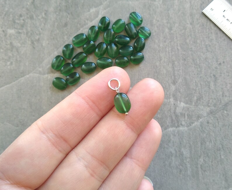 May include: A close-up of a green oval-shaped pendant with a silver bail. The pendant is held in a hand, and there are many other green oval-shaped beads in the background.