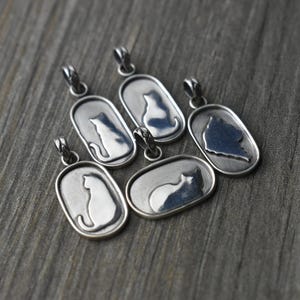 May include: Silver-toned cat pendant necklaces. Each pendant features a cat silhouette within an oval frame. The pendants are displayed on a weathered, gray wooden surface.