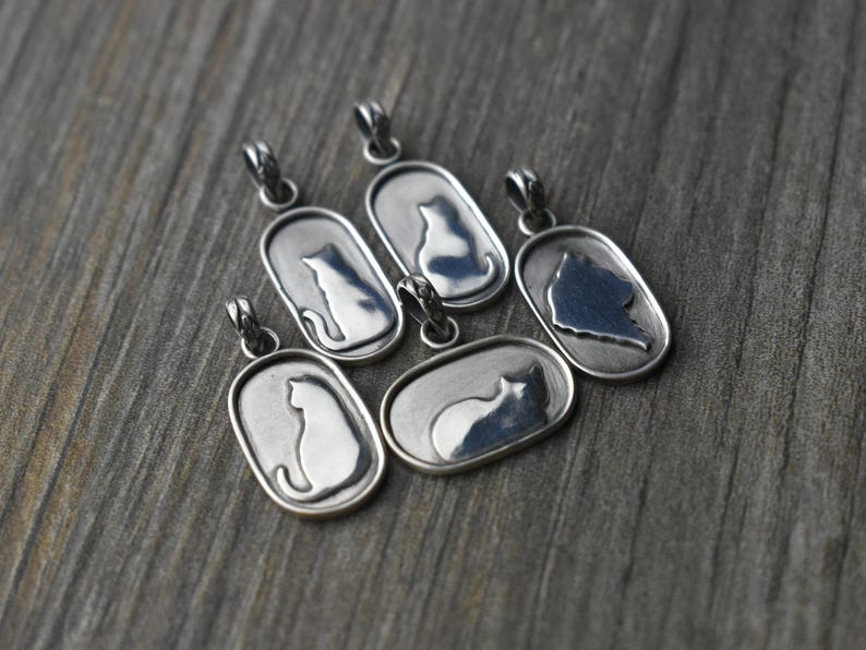 May include: Silver-toned cat pendant necklaces. Each pendant features a cat silhouette within an oval frame. The pendants are displayed on a weathered, gray wooden surface.