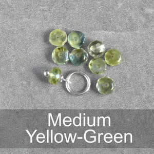 Op de afbeelding: Een verzameling geelgroene edelstenen, sommige met een lichte blauwe tint, gerangschikt op een grijs oppervlak. De edelstenen zijn rond en gefacetteerd. De tekst "Medium Yellow-Green" wordt weergegeven onder de edelstenen.