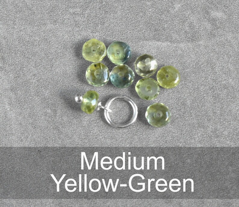 Op de afbeelding: Een verzameling geelgroene edelstenen, sommige met een lichte blauwe tint, gerangschikt op een grijs oppervlak. De edelstenen zijn rond en gefacetteerd. De tekst "Medium Yellow-Green" wordt weergegeven onder de edelstenen.