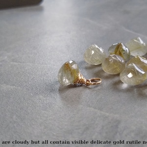May include: A group of cloudy, faceted, clear quartz stones with visible gold rutile needles. The stones are scattered on a gray surface. One stone is attached to a gold wire loop.