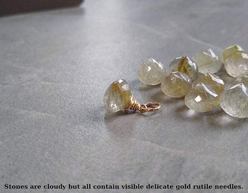 May include: A group of cloudy, faceted, clear quartz stones with visible gold rutile needles. The stones are scattered on a gray surface. One stone is attached to a gold wire loop.
