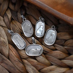 May include: A collection of silver-toned cat pendant necklaces. Each pendant features a cat silhouette within an oval or rectangular frame. The pendants are displayed on a woven, brown surface.