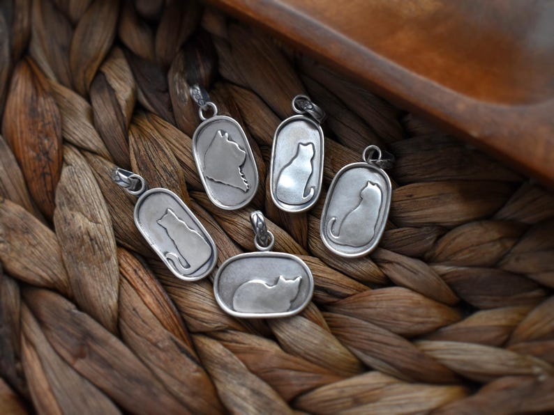 May include: A collection of silver-toned cat pendant necklaces. Each pendant features a cat silhouette within an oval or rectangular frame. The pendants are displayed on a woven, brown surface.