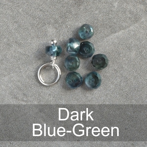Op de afbeelding: Een close-up van donkerblauwgroene gefacetteerde glazen kralen met een zilveren springring. De kralen zijn in een halve cirkel gerangschikt met de springring aan de bovenkant.