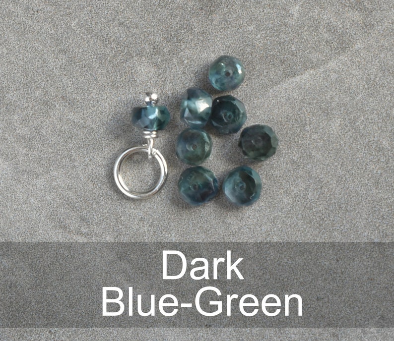 Op de afbeelding: Een close-up van donkerblauwgroene gefacetteerde glazen kralen met een zilveren springring. De kralen zijn in een halve cirkel gerangschikt met de springring aan de bovenkant.
