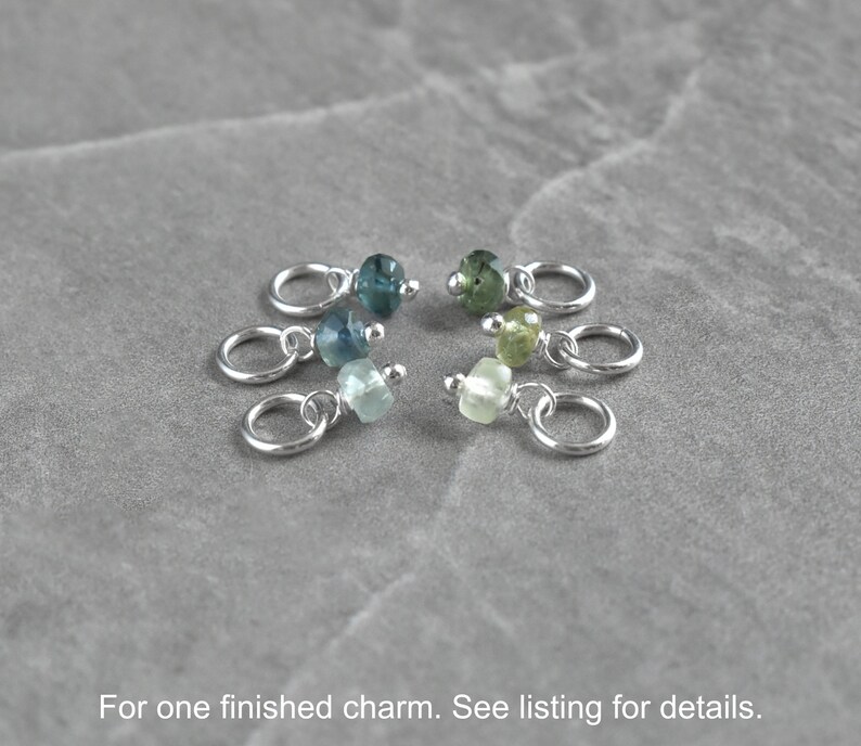 Op de afbeelding: Zes zilveren bedels met verschillende gekleurde edelsteen kralen. De kralen zijn in tinten blauw en groen. De bedels zijn voor het maken van sieraden.