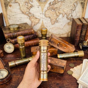 May include: A cylindrical brass telescope featuring an engraved label that reads Gilbert & Sons, London, 1917. The handheld device has a polished metallic finish with decorative bands and a circular eyepiece, showcasing a vintage nautical design.