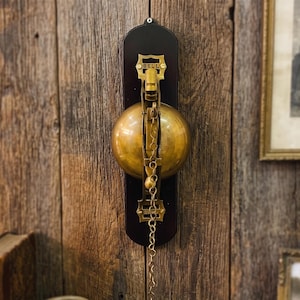 May include: A wall-mounted bell featuring a polished brass dome and a dark brown wooden backplate. The brass mechanism includes a vertical bracket with the word BELL embossed at the top and a long metal pull chain hanging from the center.