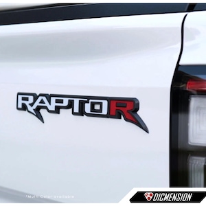以下が含まれることがあります： 白の車に取り付けられた、黒、白、赤の「RAPTOR」エンブレム。エンブレムは、黒いアウトラインと赤い「R」を持つ様式化されたフォントが特徴です。画像は、車両のボディのクローズアップです。