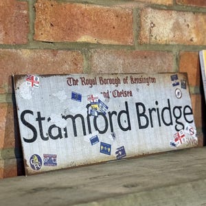 May include: A weathered metal sign reading "Stamford Bridge" with the text "The Royal Borough of Kensington and Chelsea". The sign is decorated with stickers and sits on a wooden shelf against a brick wall.